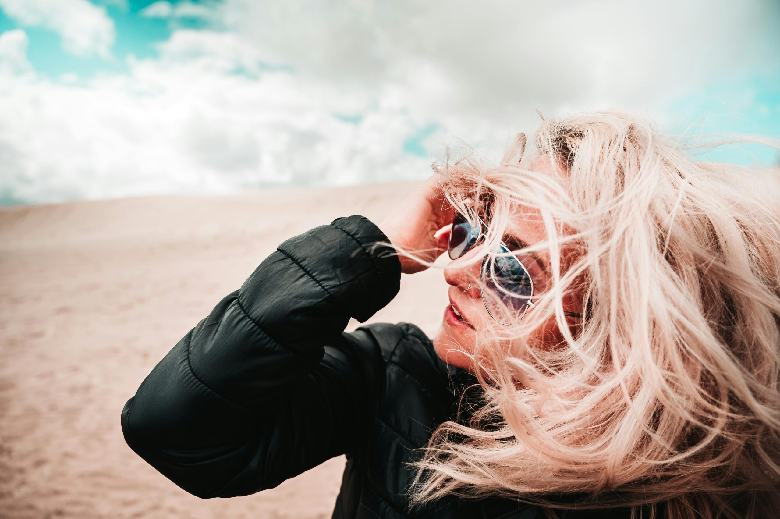 A woman with windswept hair in sunglasses enjoys a breezy day on a sandy beach.