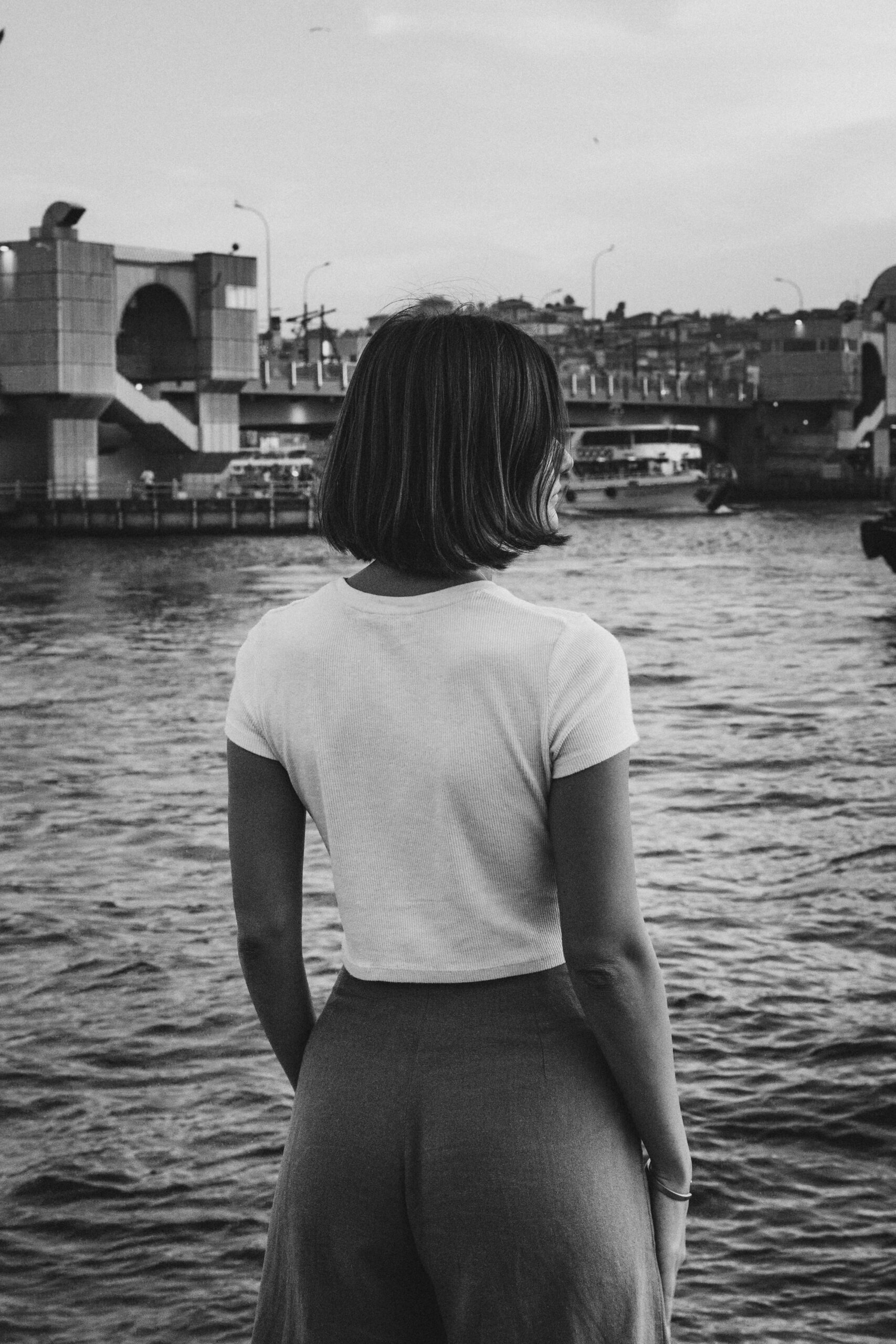 Woman with short hair standing by a river, overlooking urban architecture in black and white.